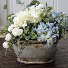 white and blue flowers are in an old metal pot on a table with greenery