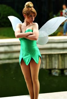 a woman dressed as tinkerbell standing in front of a pond with her arms crossed