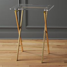 a clear glass and gold metal desk with wooden legs on the floor in front of a gray wall