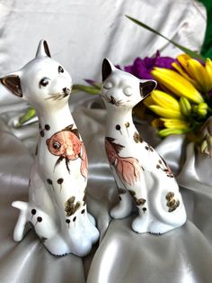 two ceramic cats sitting next to each other on a white cloth with flowers in the background