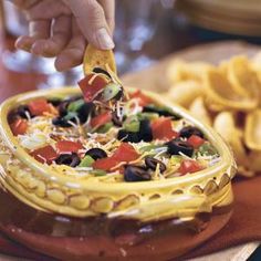 a person dipping a tortilla into a bowl filled with black olives, peppers and cheese