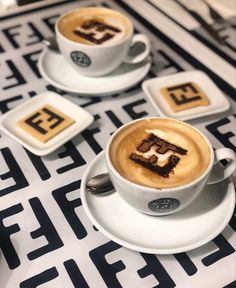 two cups of cappuccino with the letter e on them sitting on a black and white tablecloth