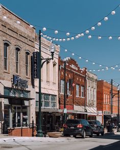 cars are parked on the street in front of buildings with lights strung over them and people walking down the sidewalk