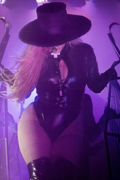 a woman with long blonde hair wearing a black outfit and hat sitting on a stage