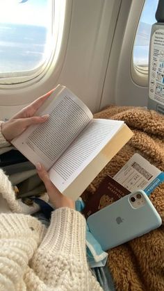 a woman reading a book while sitting on an airplane with her cell phone and passport