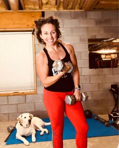 a woman in red pants and black tank top standing next to a white dog on a blue mat