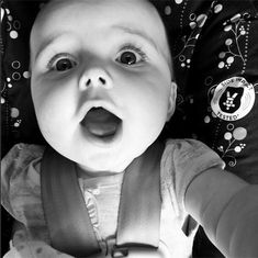 a black and white photo of a baby in a car seat with its tongue out