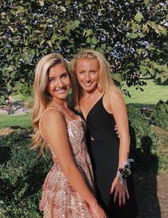 two beautiful women standing next to each other in front of a green tree and bushes