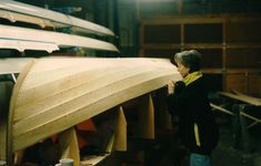 a young boy standing next to a wooden boat in a garage with other boats behind him