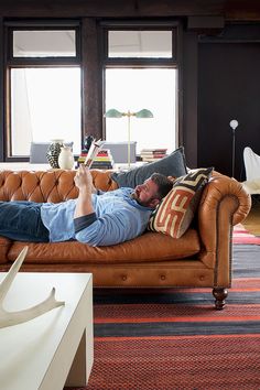 a man laying on top of a brown couch in a living room next to two windows