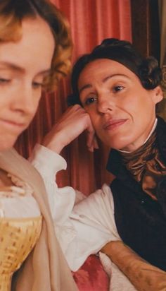 two women dressed in period clothing sitting next to each other and one is touching her ear