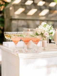 three glasses filled with pink liquid sitting on top of a white table next to candles and flowers