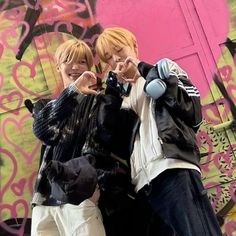 two young men standing next to each other in front of a graffiti covered wall with one pointing at the camera