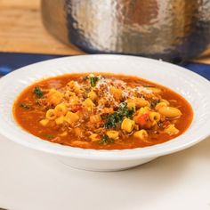 a white bowl filled with pasta soup on top of a wooden table next to a silver pot