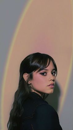 a woman with long black hair standing in front of a rainbow colored wall and looking at the camera