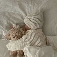 a small teddy bear laying on top of a bed next to a white blanket and hat