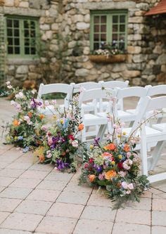 rows of white folding chairs with colorful flowers on the back set up for an outdoor ceremony