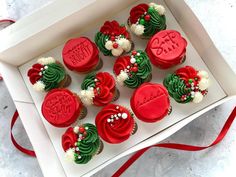 twelve decorated christmas cupcakes in a white box with red ribbon on the side