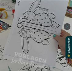 a person holding up a coloring book in front of a table with drawings on it