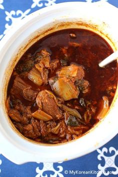 a white bowl filled with stew and meat on top of a blue table cloth next to a spoon