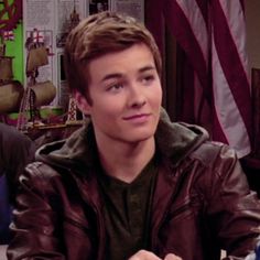 a young man sitting at a table with an american flag in the background and another person standing behind him