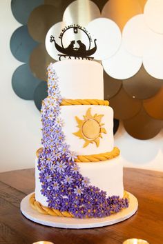 a three tiered cake decorated with purple flowers and a sun decoration on the top