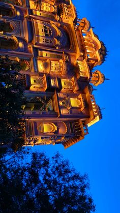 an ornate building lit up at night with trees in the foreground and blue sky behind it