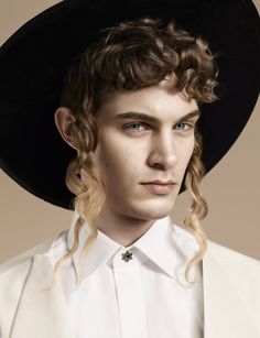 a young man with long hair wearing a black hat and white shirt, standing in front of a beige background