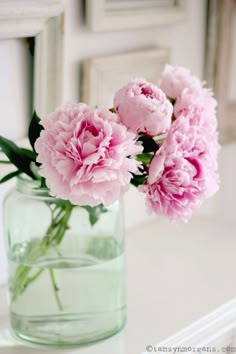 some pink flowers are in a glass vase on a white counter top with an antique frame behind them