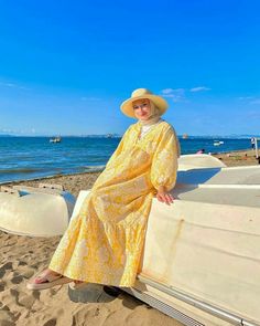 a woman in a yellow dress and hat sitting on a boat at the beach by the water