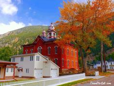 an old red church in the fall with trees and houses around it, on a sunny day