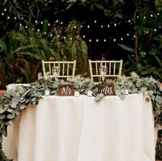 the table is set up with greenery and mr and mrs signs on it, along with two chairs