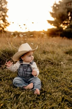 a baby sitting in the grass wearing a cowboy hat and looking up at the sky