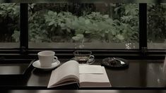 an open book, coffee cup and saucer sit on a table in front of a window