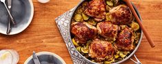 a pan filled with chicken and vegetables on top of a wooden table next to utensils