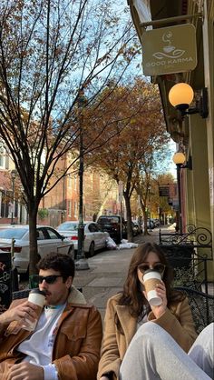 two people sitting on a bench drinking coffee and talking to each other in the street