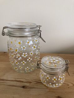 two glass jars with yellow and white flowers on them sitting on a wooden table next to each other