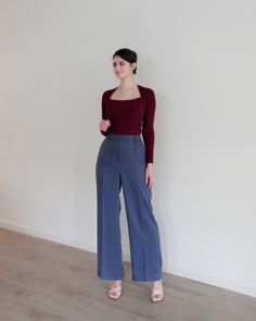 a woman standing in front of a white wall wearing blue pants and a maroon top