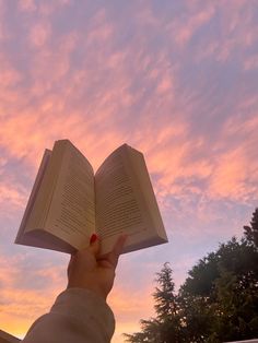 a person is holding an open book in front of the sky with trees and clouds