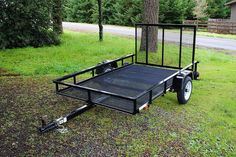 a black trailer is parked in the grass near a tree and some trees on the side of the road