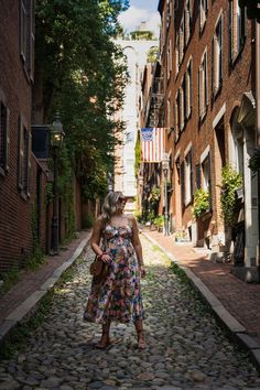 a woman in a floral dress walking down a cobblestone street with buildings on both sides