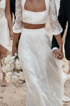 a bride and groom holding hands walking on the beach with other people in the background