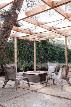 a fire pit surrounded by chairs under a wooden pergolan roof with trees and bushes in the background