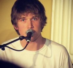 a young man singing into a microphone in front of a yellow wall and white curtains