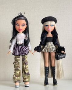 two dolls standing next to each other in front of a white wall, one holding a purse