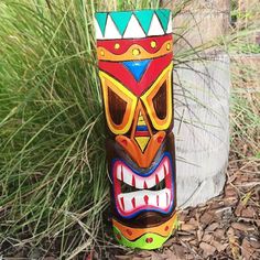 a colorful tiki mask sitting on the ground next to some grass and rocks with plants in the background