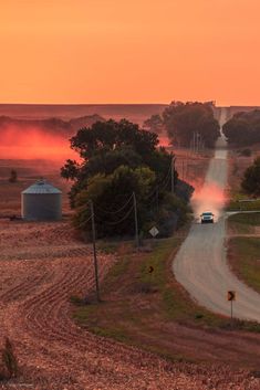 a truck driving down a dirt road next to a field at sunset in the country