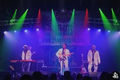 three men on stage with green and red lights in the background, one man is playing guitar