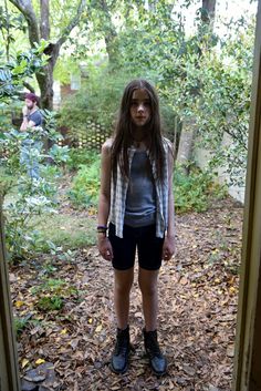 a girl standing in front of a door with leaves on the ground and trees behind her