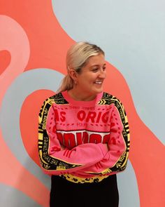 a woman standing in front of a wall wearing a pink sweater with an origi design on it